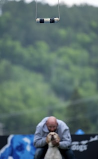 A trapeze handle hangs suspended in the air with a blurred background of lush green foliage. Below, a person is crouched, embracing a small light-colored dog, while the focus remains on the suspended handle.