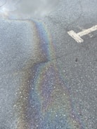 A rough asphalt surface with an iridescent oil spill reflecting a spectrum of colors. A large, white arrow indicating direction is partially visible on the pavement.