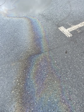 A rough asphalt surface with an iridescent oil spill reflecting a spectrum of colors. A large, white arrow indicating direction is partially visible on the pavement.