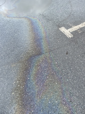 A rough asphalt surface with an iridescent oil spill reflecting a spectrum of colors. A large, white arrow indicating direction is partially visible on the pavement.