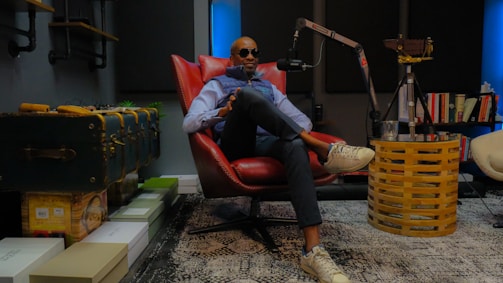 A person wearing sunglasses is sitting comfortably in a red leather chair in a cozy, modern room with a podcast setup. The room features a microphone on a stand, bookshelves, and a patterned rug. The atmosphere is sophisticated and relaxed.