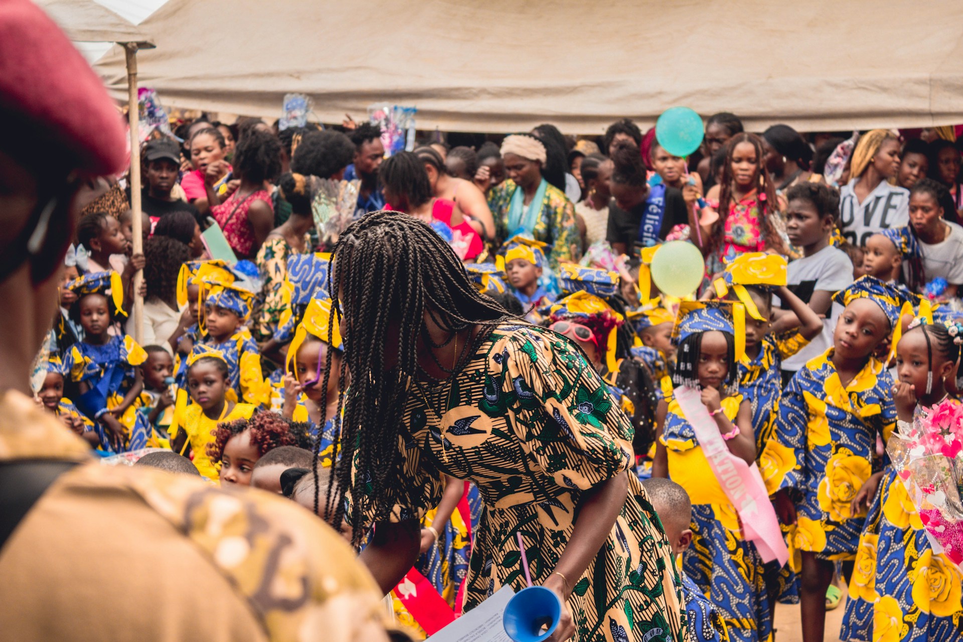 A lively gathering with many children and adults dressed in colorful traditional clothing. The children are wearing vibrant outfits with blue, yellow, and white patterns, and some have sashes. The scene appears festive with many people holding balloons and flowers.