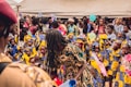 A lively gathering with many children and adults dressed in colorful traditional clothing. The children are wearing vibrant outfits with blue, yellow, and white patterns, and some have sashes. The scene appears festive with many people holding balloons and flowers.