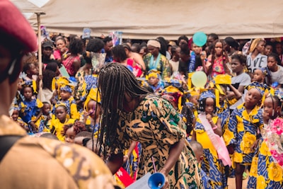 A lively gathering with many children and adults dressed in colorful traditional clothing. The children are wearing vibrant outfits with blue, yellow, and white patterns, and some have sashes. The scene appears festive with many people holding balloons and flowers.