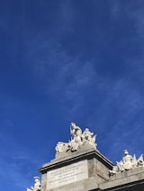 A detailed stone sculpture featuring several human figures atop a grand architectural structure. The figures are intricately carved and arranged in a classical style. The structure appears to be part of a monument with an inscription in Latin on a rectangular plaque. The sky in the background is clear and blue, adding contrast to the white sculptures.