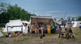 A medieval market scene with several tents set up on a grassy field. Various historical artifacts such as shields, helmets, and weapons are displayed for sale outside the tents. People dressed in period costumes are browsing the items, creating an atmosphere reminiscent of a historical reenactment. In the background, lush green trees border the scene under a clear blue sky.