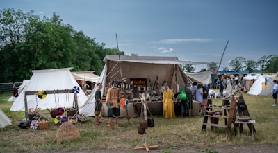 A medieval market scene with several tents set up on a grassy field. Various historical artifacts such as shields, helmets, and weapons are displayed for sale outside the tents. People dressed in period costumes are browsing the items, creating an atmosphere reminiscent of a historical reenactment. In the background, lush green trees border the scene under a clear blue sky.