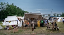 A medieval market scene with several tents set up on a grassy field. Various historical artifacts such as shields, helmets, and weapons are displayed for sale outside the tents. People dressed in period costumes are browsing the items, creating an atmosphere reminiscent of a historical reenactment. In the background, lush green trees border the scene under a clear blue sky.