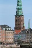 A historic cityscape features a prominent copper-topped tower in an architectural style with intricate detailing. This structure is surrounded by traditional European buildings with gabled roofs and multiple windows. A statue sits in the foreground, and people can be seen relaxing on the benches near the water's edge.