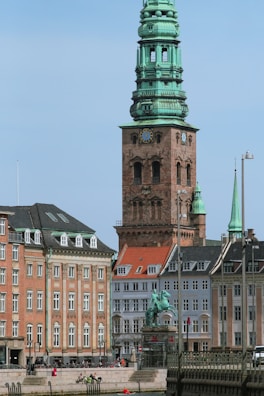 A historic cityscape features a prominent copper-topped tower in an architectural style with intricate detailing. This structure is surrounded by traditional European buildings with gabled roofs and multiple windows. A statue sits in the foreground, and people can be seen relaxing on the benches near the water's edge.
