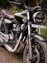 A close-up view of a dark green motorcycle with shiny chrome features and a headlight. The vehicle is parked on a dirt path, displaying a rugged and slightly muddy appearance. The brand logo is visible on the gas tank, emphasizing its make. The number plate is prominently displayed, adding to its identification.