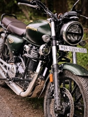 A close-up view of a dark green motorcycle with shiny chrome features and a headlight. The vehicle is parked on a dirt path, displaying a rugged and slightly muddy appearance. The brand logo is visible on the gas tank, emphasizing its make. The number plate is prominently displayed, adding to its identification.