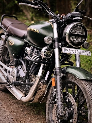 A close-up view of a dark green motorcycle with shiny chrome features and a headlight. The vehicle is parked on a dirt path, displaying a rugged and slightly muddy appearance. The brand logo is visible on the gas tank, emphasizing its make. The number plate is prominently displayed, adding to its identification.