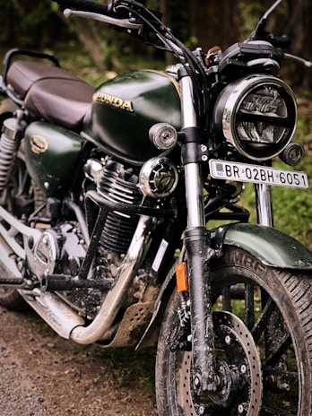 A close-up view of a dark green motorcycle with shiny chrome features and a headlight. The vehicle is parked on a dirt path, displaying a rugged and slightly muddy appearance. The brand logo is visible on the gas tank, emphasizing its make. The number plate is prominently displayed, adding to its identification.
