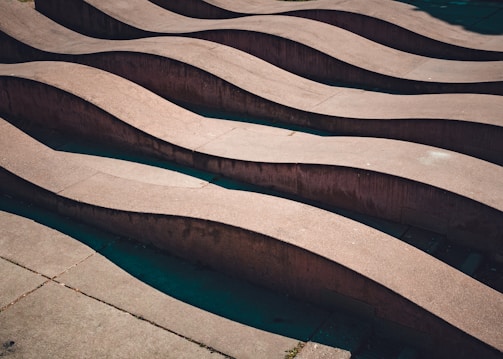 A series of concrete waves or undulating structures create a rhythmic pattern. The light casts soft shadows, enhancing the depth and curvature of the formations. The texture of the surface is smooth, and the undulations extend across a flat surface, suggesting an architectural or artistic installation.