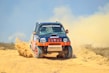 A rugged off-road vehicle with red and black accents speeds through a sandy terrain, kicking up a cloud of dust behind it. The vehicle's design features sponsor logos and vibrant orange wheels, reflecting its participation in a rally or off-road race.