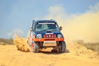 A rugged off-road vehicle with red and black accents speeds through a sandy terrain, kicking up a cloud of dust behind it. The vehicle's design features sponsor logos and vibrant orange wheels, reflecting its participation in a rally or off-road race.