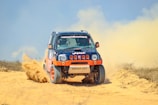 A rugged off-road vehicle with red and black accents speeds through a sandy terrain, kicking up a cloud of dust behind it. The vehicle's design features sponsor logos and vibrant orange wheels, reflecting its participation in a rally or off-road race.