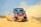A rugged off-road vehicle with red and black accents speeds through a sandy terrain, kicking up a cloud of dust behind it. The vehicle's design features sponsor logos and vibrant orange wheels, reflecting its participation in a rally or off-road race.