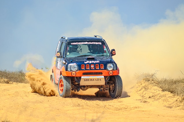 A rugged off-road vehicle with red and black accents speeds through a sandy terrain, kicking up a cloud of dust behind it. The vehicle's design features sponsor logos and vibrant orange wheels, reflecting its participation in a rally or off-road race.