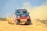 A rugged off-road vehicle with red and black accents speeds through a sandy terrain, kicking up a cloud of dust behind it. The vehicle's design features sponsor logos and vibrant orange wheels, reflecting its participation in a rally or off-road race.
