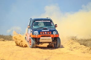 A rugged off-road vehicle with red and black accents speeds through a sandy terrain, kicking up a cloud of dust behind it. The vehicle's design features sponsor logos and vibrant orange wheels, reflecting its participation in a rally or off-road race.