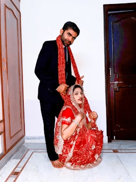 A man in a formal black suit stands next to a woman dressed in a traditional bright red and gold embroidered outfit. She is crouching on the floor with a dupatta draped over her head. The setting appears to be indoors with a tiled floor and wooden doors.