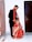 A man in a formal black suit stands next to a woman dressed in a traditional bright red and gold embroidered outfit. She is crouching on the floor with a dupatta draped over her head. The setting appears to be indoors with a tiled floor and wooden doors.