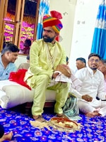 A man dressed in traditional attire, including a colorful turban and a string of pearls, sits on a cushion in a room with several other people. The room features vibrant blue and white curtains and a decorated blue carpet with floral patterns. Some people are sitting cross-legged, engaging in what appears to be a ceremonial or cultural event.
