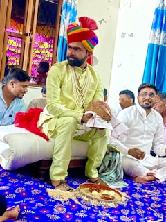 A man dressed in traditional attire, including a colorful turban and a string of pearls, sits on a cushion in a room with several other people. The room features vibrant blue and white curtains and a decorated blue carpet with floral patterns. Some people are sitting cross-legged, engaging in what appears to be a ceremonial or cultural event.