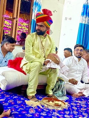 A man dressed in traditional attire, including a colorful turban and a string of pearls, sits on a cushion in a room with several other people. The room features vibrant blue and white curtains and a decorated blue carpet with floral patterns. Some people are sitting cross-legged, engaging in what appears to be a ceremonial or cultural event.