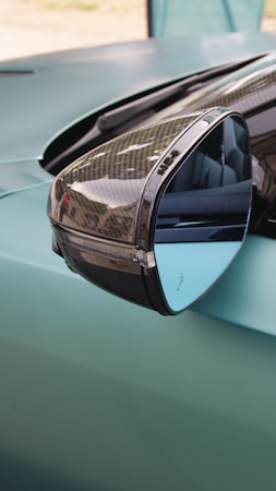 A close-up view of a car's side mirror, reflecting its sleek design. The mirror has a glossy, slightly curved surface with reflections of the surroundings. The car is painted in a muted turquoise shade, and the mirror casing appears to have a carbon fiber texture.