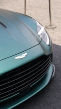 A close-up of the front section of a luxury car, featuring a sleek, metallic green finish. The prominent grille and badge add a sense of sophistication. The headlight design is modern and sharp, reflecting light. The car is positioned on a textured surface, with metallic stanchions visible in the background.