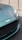 A close-up of the front section of a luxury car, featuring a sleek, metallic green finish. The prominent grille and badge add a sense of sophistication. The headlight design is modern and sharp, reflecting light. The car is positioned on a textured surface, with metallic stanchions visible in the background.