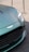 A close-up of the front section of a luxury car, featuring a sleek, metallic green finish. The prominent grille and badge add a sense of sophistication. The headlight design is modern and sharp, reflecting light. The car is positioned on a textured surface, with metallic stanchions visible in the background.