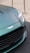 A close-up of the front section of a luxury car, featuring a sleek, metallic green finish. The prominent grille and badge add a sense of sophistication. The headlight design is modern and sharp, reflecting light. The car is positioned on a textured surface, with metallic stanchions visible in the background.