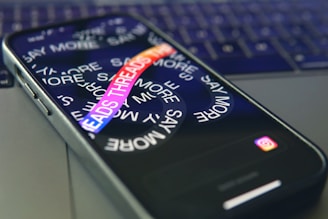 A smartphone lies on a laptop keyboard with the screen displaying swirling text that reads 'SAY MORE' and 'THREADS' in colorful letters. The Instagram logo is visible at the bottom of the phone's screen.