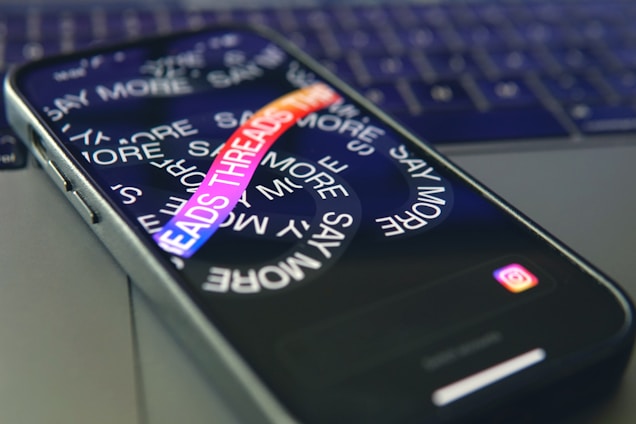 A smartphone lies on a laptop keyboard with the screen displaying swirling text that reads 'SAY MORE' and 'THREADS' in colorful letters. The Instagram logo is visible at the bottom of the phone's screen.