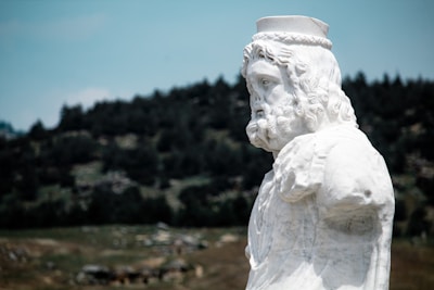 A marble statue of a bearded man is set against a blurred background of a rugged landscape with sparse vegetation and a mountain range. The statue, featuring intricate details, appears to depict a historical or mythological figure, wearing a headband or crown.