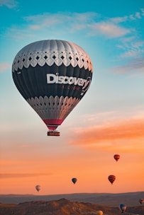 A large hot air balloon dominates the sky, featuring a decorative pattern and the word 'Discovery'. The backdrop reveals a serene landscape with a series of other colorful balloons floating gently against a gradient sky, transitioning from blue at the top to warm orange hues near the horizon.