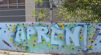 A colorful graffiti mural on a wall with the word 'APREN!' in large, bold letters. Surrounding the text are colorful spray paint dots in hues of blue, yellow, purple, and green. There are pencil illustrations on either side of the text, emphasizing a learning theme. Behind the wall is a metal fence and some greenery.