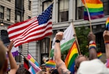 A lively street scene with a crowd of people holding various flags, including an American flag and multiple rainbow flags. The setting is urban, with tall buildings as a backdrop. Some individuals are wearing colorful wristbands and waving their hands in celebration.