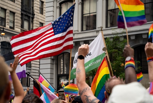 A lively street scene with a crowd of people holding various flags, including an American flag and multiple rainbow flags. The setting is urban, with tall buildings as a backdrop. Some individuals are wearing colorful wristbands and waving their hands in celebration.