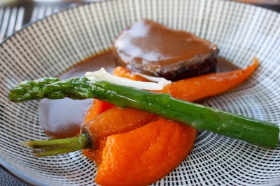 A gourmet dish featuring a piece of cooked meat drizzled with a rich brown sauce, accompanied by vibrant orange carrot puree, a whole cooked carrot, and a steamed green asparagus spear. The food is elegantly arranged on a patterned plate, showcasing a blend of colors and textures.