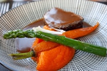 A gourmet dish featuring a piece of cooked meat drizzled with a rich brown sauce, accompanied by vibrant orange carrot puree, a whole cooked carrot, and a steamed green asparagus spear. The food is elegantly arranged on a patterned plate, showcasing a blend of colors and textures.