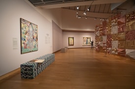 A modern art gallery with wooden flooring features several paintings on the walls and a decorative installation made of various metallic and geometric elements. A long bench with a wave pattern fabric is placed in the foreground, with an open book on it. A person is observing one of the paintings.