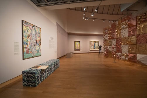 A modern art gallery with wooden flooring features several paintings on the walls and a decorative installation made of various metallic and geometric elements. A long bench with a wave pattern fabric is placed in the foreground, with an open book on it. A person is observing one of the paintings.