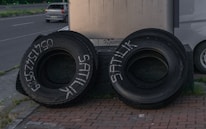 Two large tires are positioned upright on a brick-paved sidewalk. One tire has writing in white paint or chalk, displaying a phone number and the word 'SATILIK'. A parked silver car and another vehicle are partly visible behind the tires, alongside a metal structure.