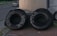 Two large tires are positioned upright on a brick-paved sidewalk. One tire has writing in white paint or chalk, displaying a phone number and the word 'SATILIK'. A parked silver car and another vehicle are partly visible behind the tires, alongside a metal structure.