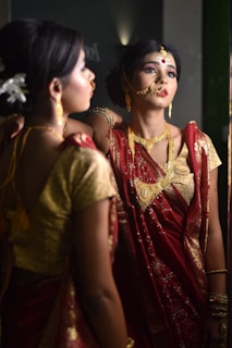 A woman adorned in traditional Indian attire is gazing at her reflection in the mirror. She wears a vibrant red saree with gold embroidery and heavy traditional jewelry, including a nose ring and a headpiece. Her makeup is meticulously done, emphasizing her eyes and lips, and she has flowers tucked into her neatly styled hair.